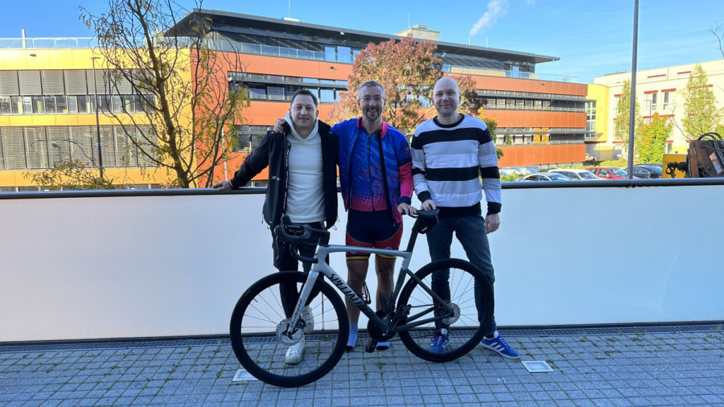 Three men stand outdoors on a sunny day with a cyclist in the center holding a bike. Behind them, bright buildings and trees add to a cheerful atmosphere.