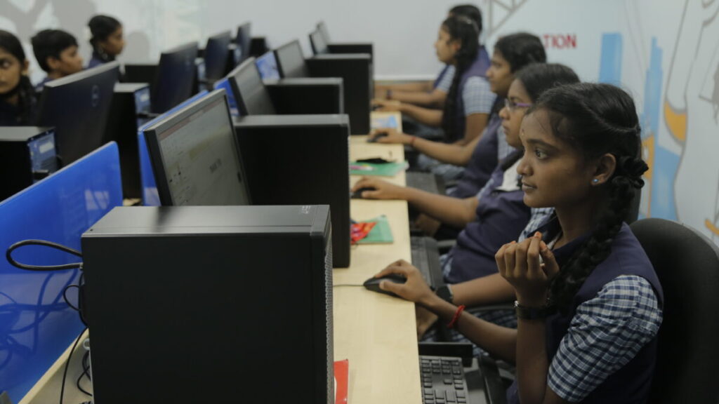 Students in school uniforms sit attentively at a row of computer workstations, focused on their monitors. The room conveys a studious atmosphere.