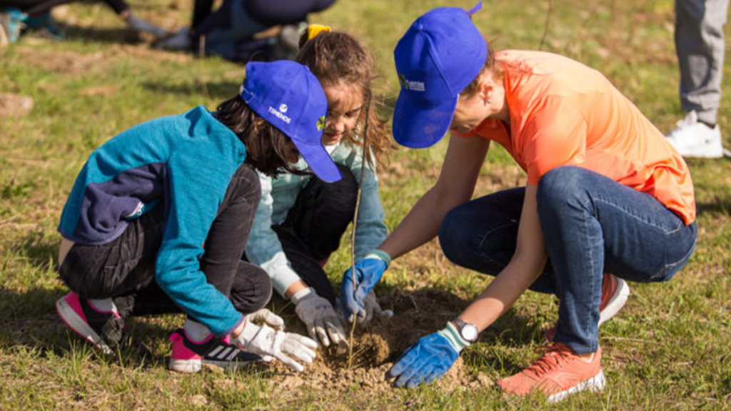 Two children and an adult, wearing blue hats and gloves, are crouched on grass planting a small tree. The scene conveys teamwork and environmental care.