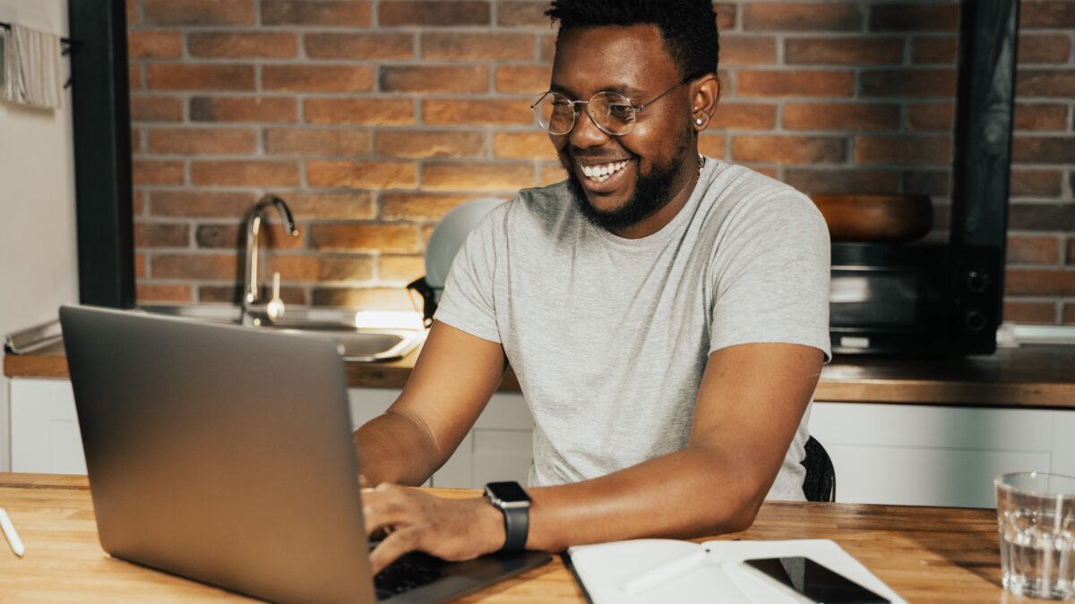 Man in glasses and a grey t-shirt smiles while using a laptop at a wooden table in a cozy, brick-walled kitchen. A notebook and phone lie next to him.