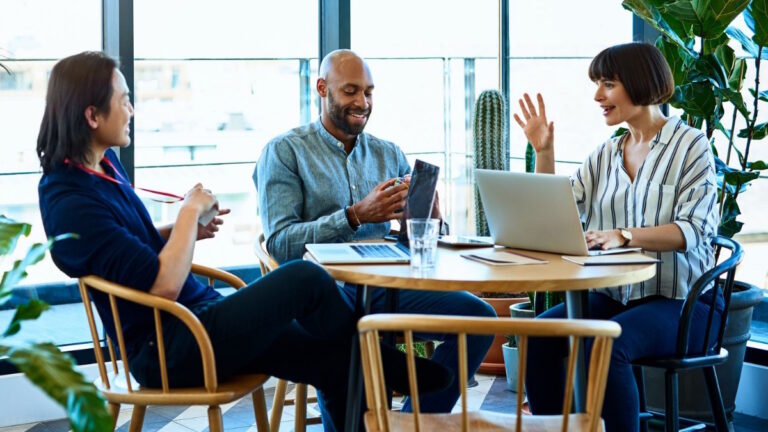 Three people sitting around a wooden table chatting. One person in the middle is on their phone. The person to the right has a laptop in front of them.
