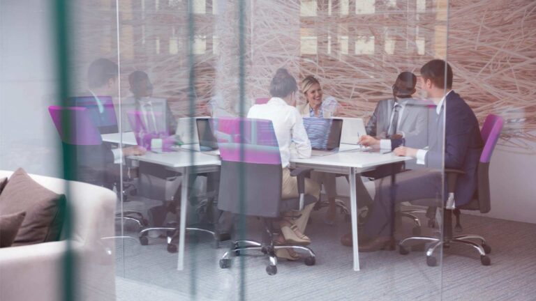 Glass walls surrounding a small office where several people are seen sitting down and collaborating.