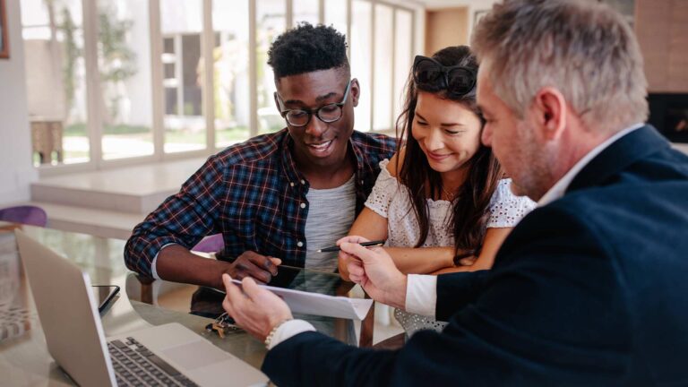 A couple is looking at a screen of a tablet being held by a businessman. He is pointinf to something on the display to draw their attention.