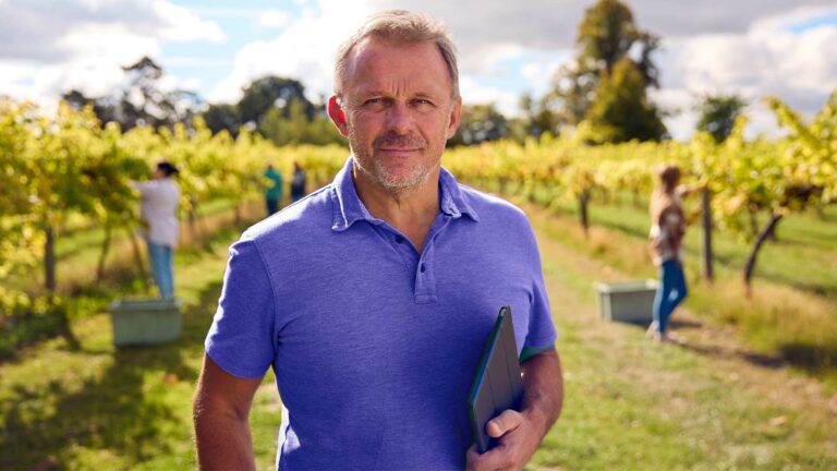 Man standing facing the camera with a tablet to his side. In the background is a farm where workers are picking fruit.