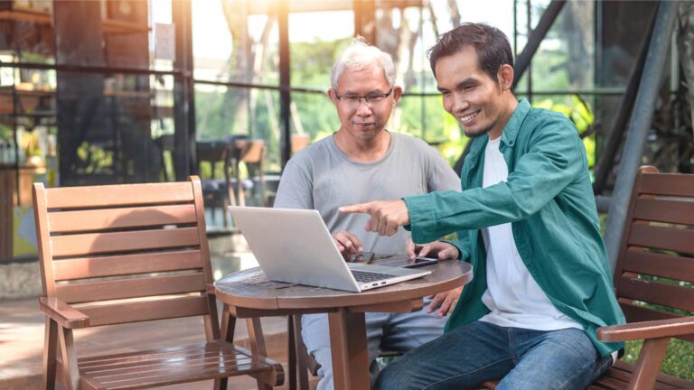 An elderly man and young man sit outside on garden furniture. The younger man is pointing to a laptop screen explaining something to the older man.