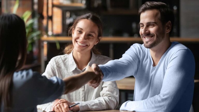 A couple is facing another person. The woman is smiling and the man is shaking hands with the other person.