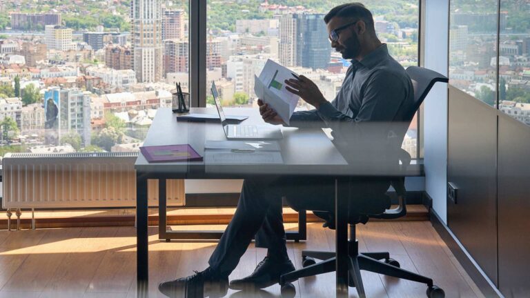 Side view of man looking at documents in his office at his desk. In the background a floor-to-ceiling window displays a city beneath.