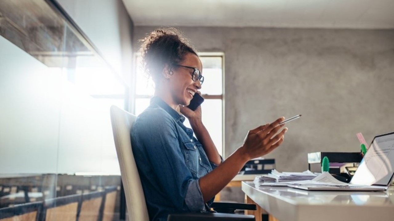 User on her mobile phone and sitting at her desk viewing her laptop.