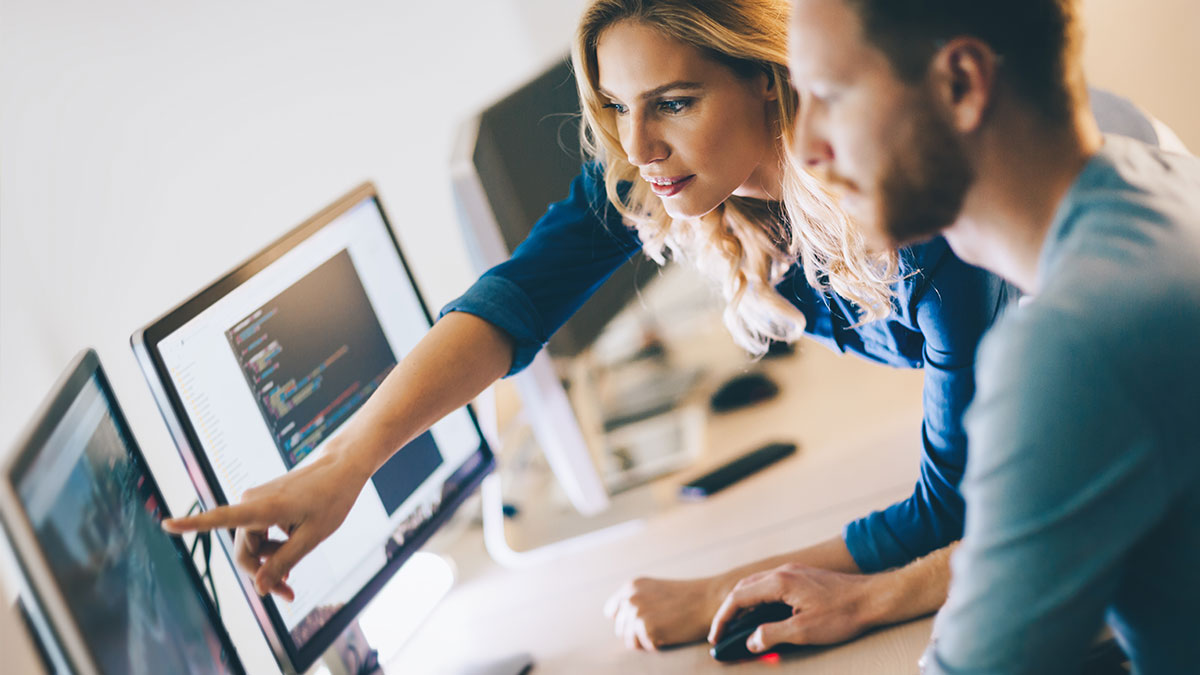 A woman and man collaborating at computers, focused and engaged. The woman points at code on a monitor, conveying teamwork and concentration.