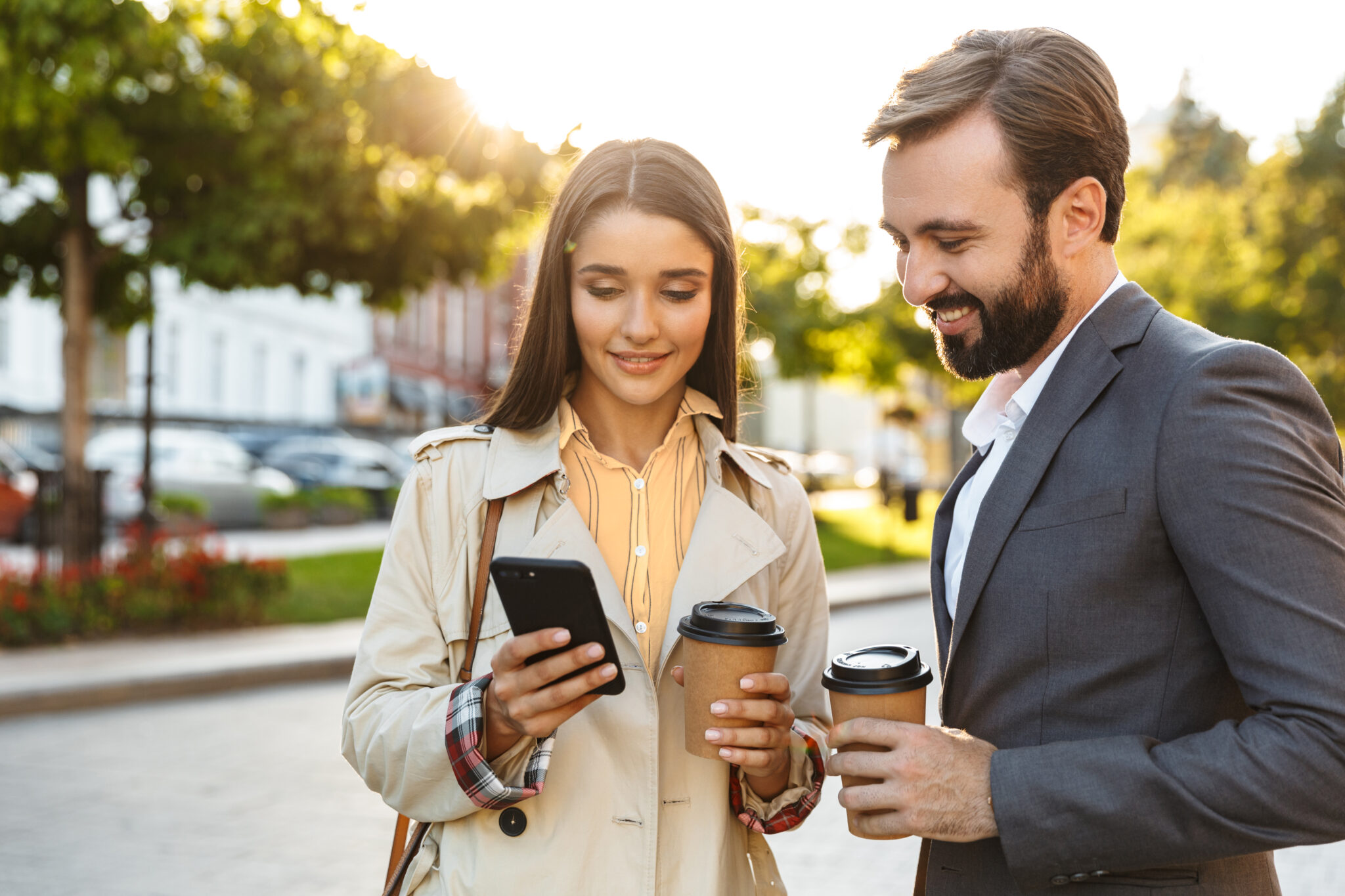 A woman and man, both smiling, stand outdoors holding coffee cups. She checks her phone while he looks on. Sunlight filters through green trees behind them.