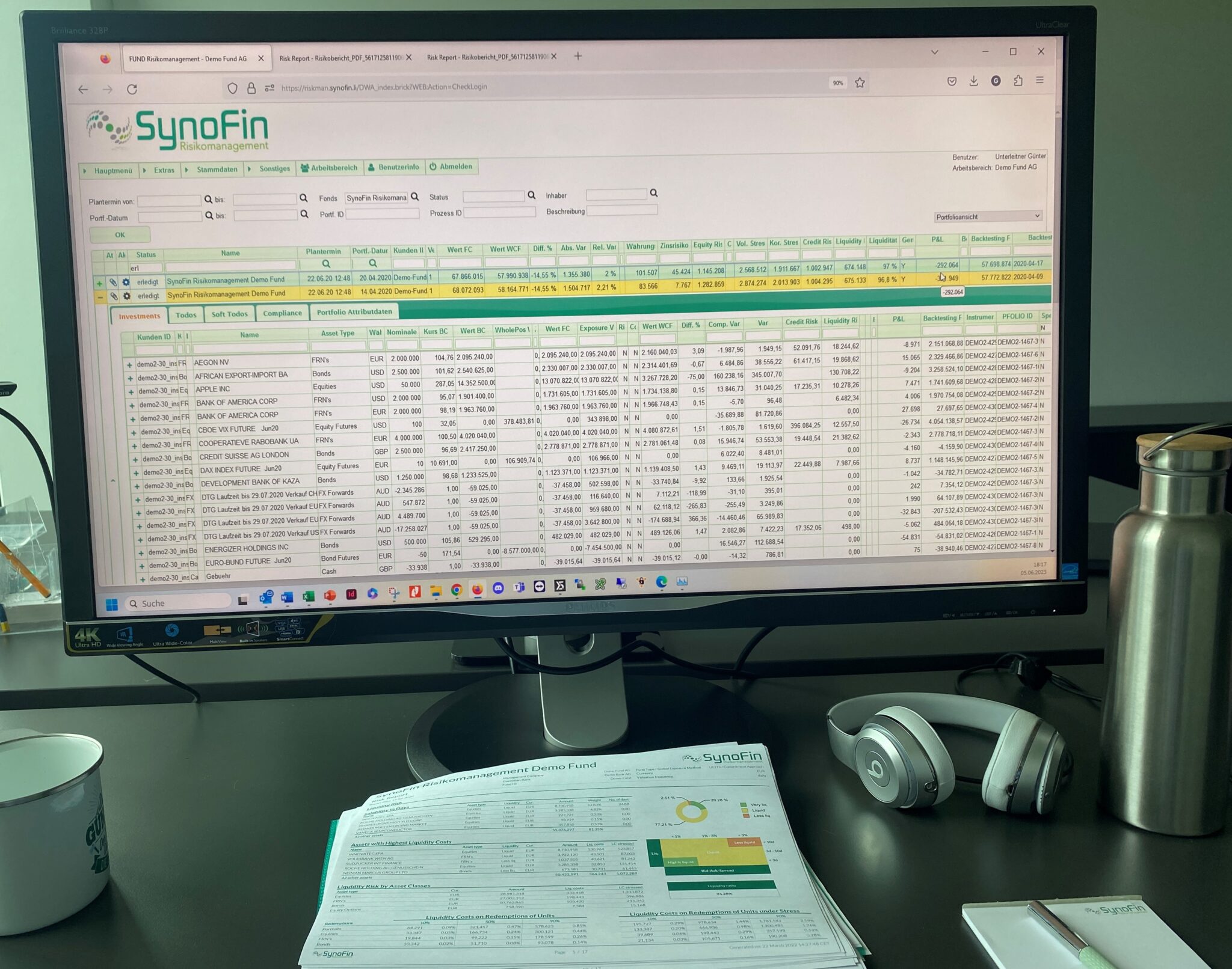 A computer monitor displays financial data on SynoFin Risk Management software. A desk has headphones, a metal bottle, a coffee mug, and documents.