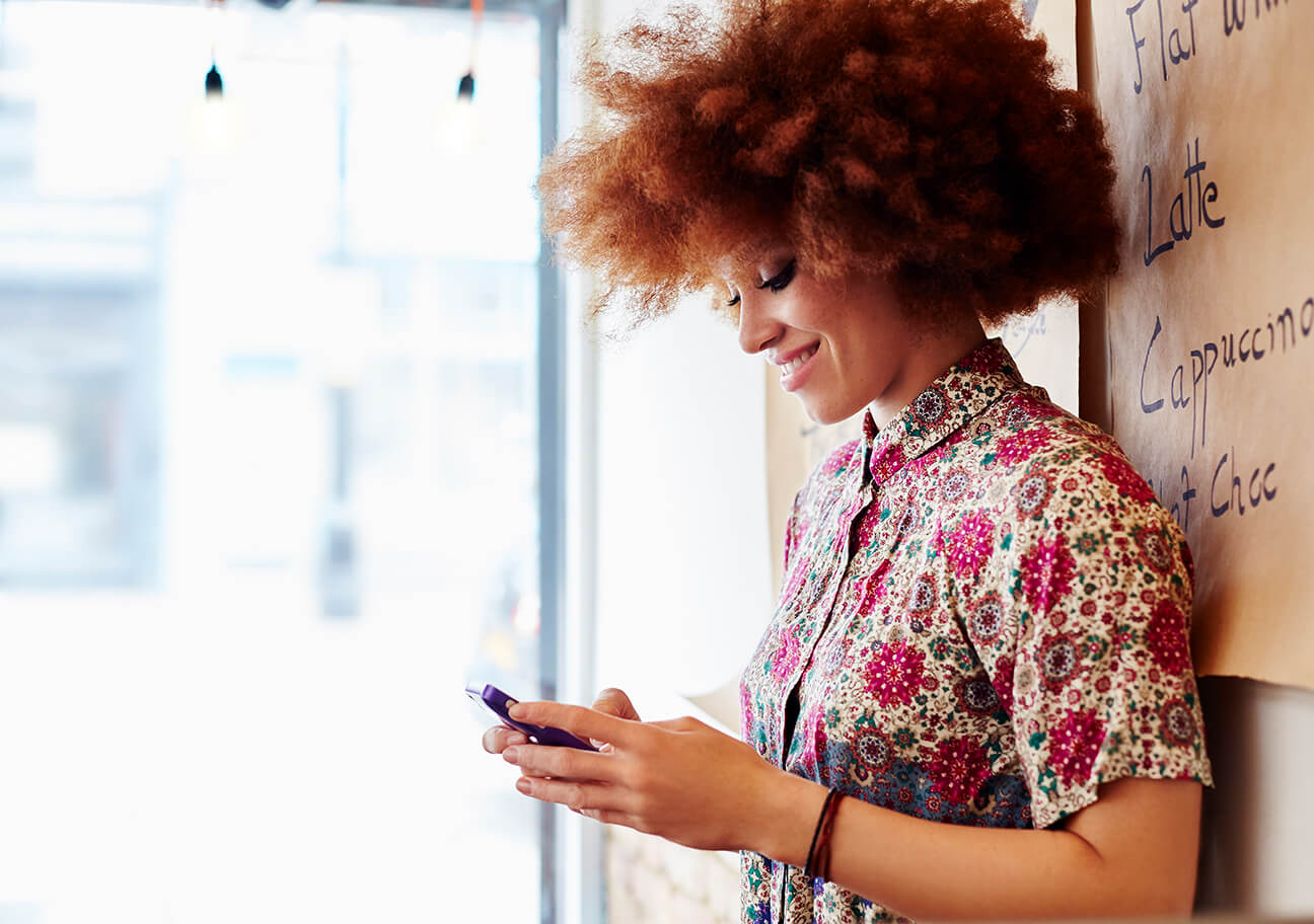 A woman with curly hair smiles while using her phone inside a cafe. She wears a colorful floral shirt. Warm light and a menu in the background add coziness.