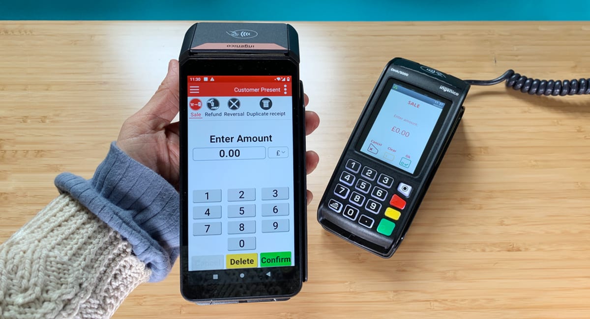 A hand holds a mobile device displaying a payment app's keypad interface. Next to it is a card reader on a wooden surface. The scene conveys a modern, easy payment process.
