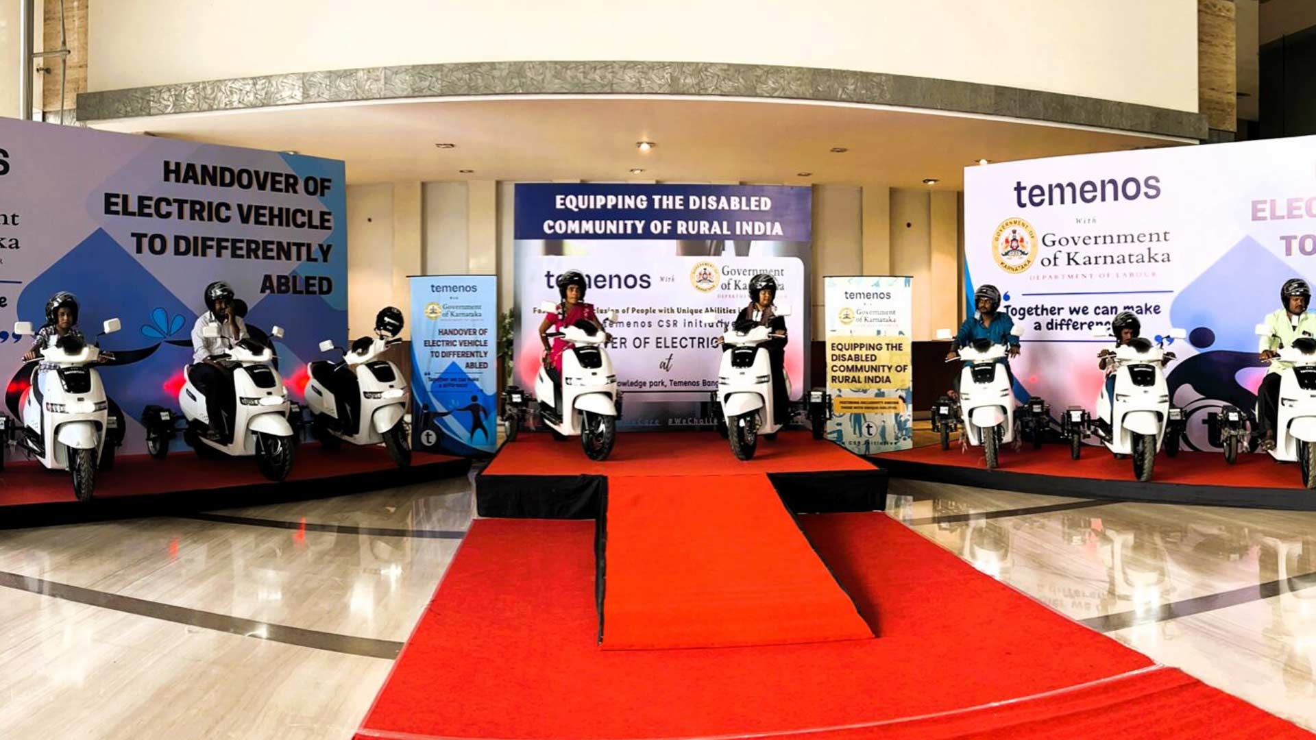 Seven individuals on white electric scooters on a stage with red carpet. Banners read "Handover of Electric Vehicle to Differently Abled." Event is organized by Temenos and the Government of Karnataka.