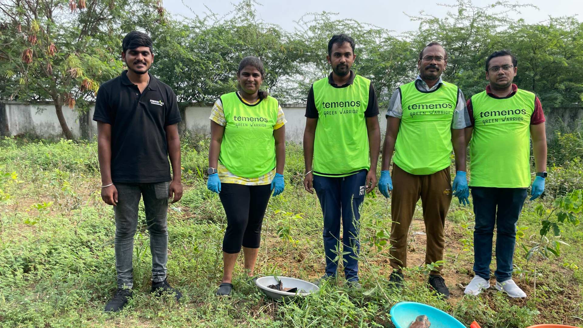 Five people are standing in a grassy area with trees in the background. Four wear bright green vests and blue gloves, suggesting a community cleaning activity.