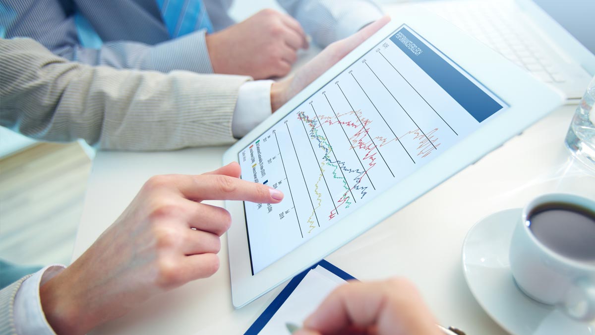 Two people discussing a tablet displaying a line graph. One points at the screen. A cup of coffee and a notepad are nearby, suggesting a business setting.
