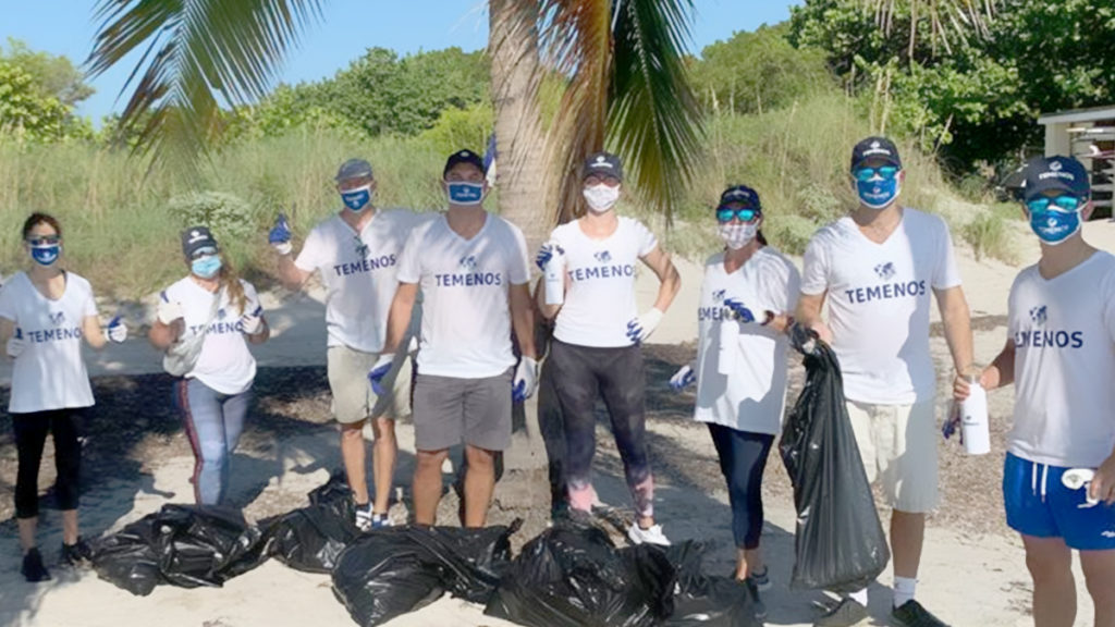 A group of people wearing white "Temenos" t-shirts stand on a beach, smiling and holding trash bags. They appear to be participating in a cleanup event.