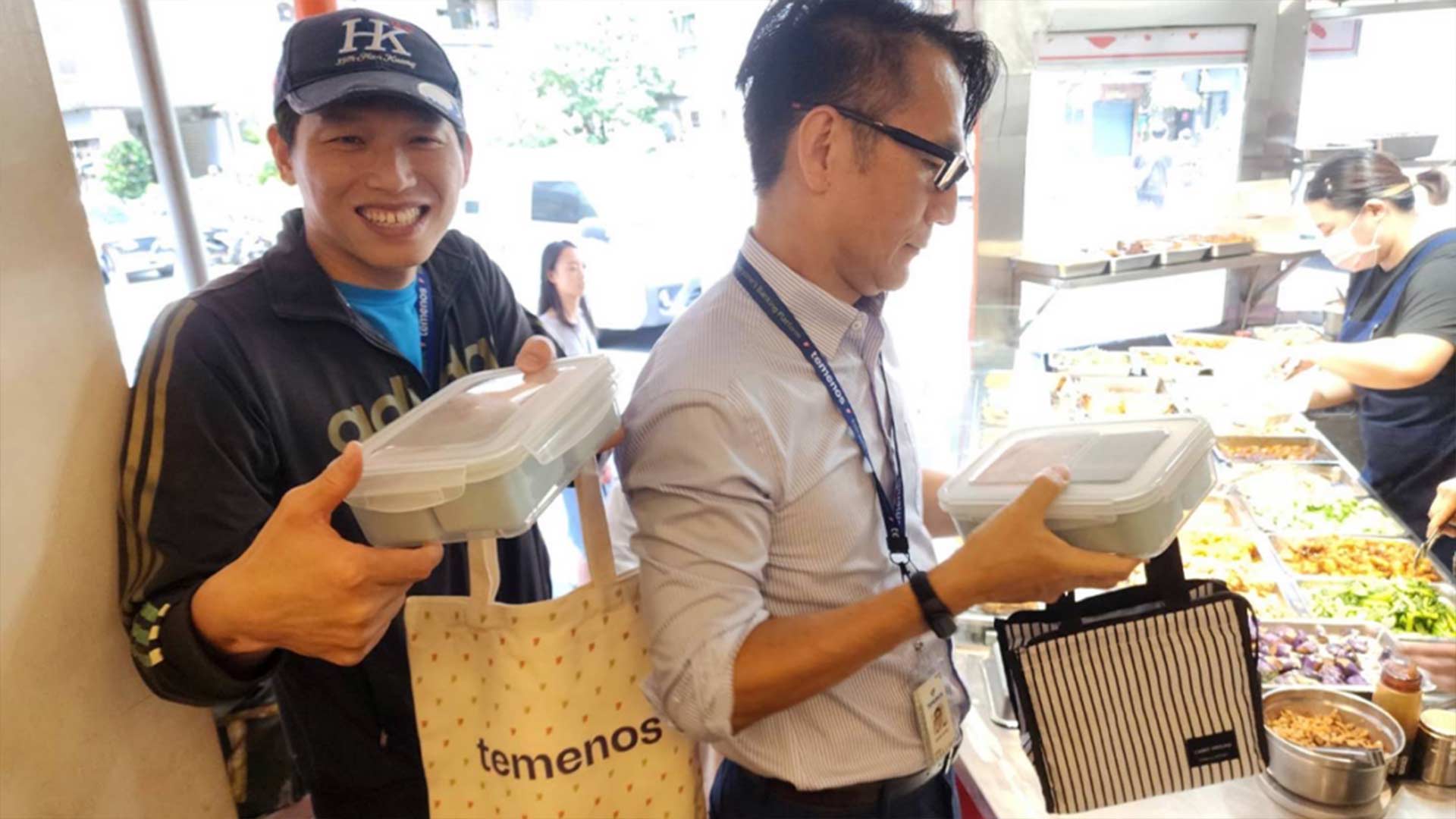 Two men smiling while holding food containers and bags in a bright restaurant. A masked server is filling dishes in the background. Casual, cheerful scene.