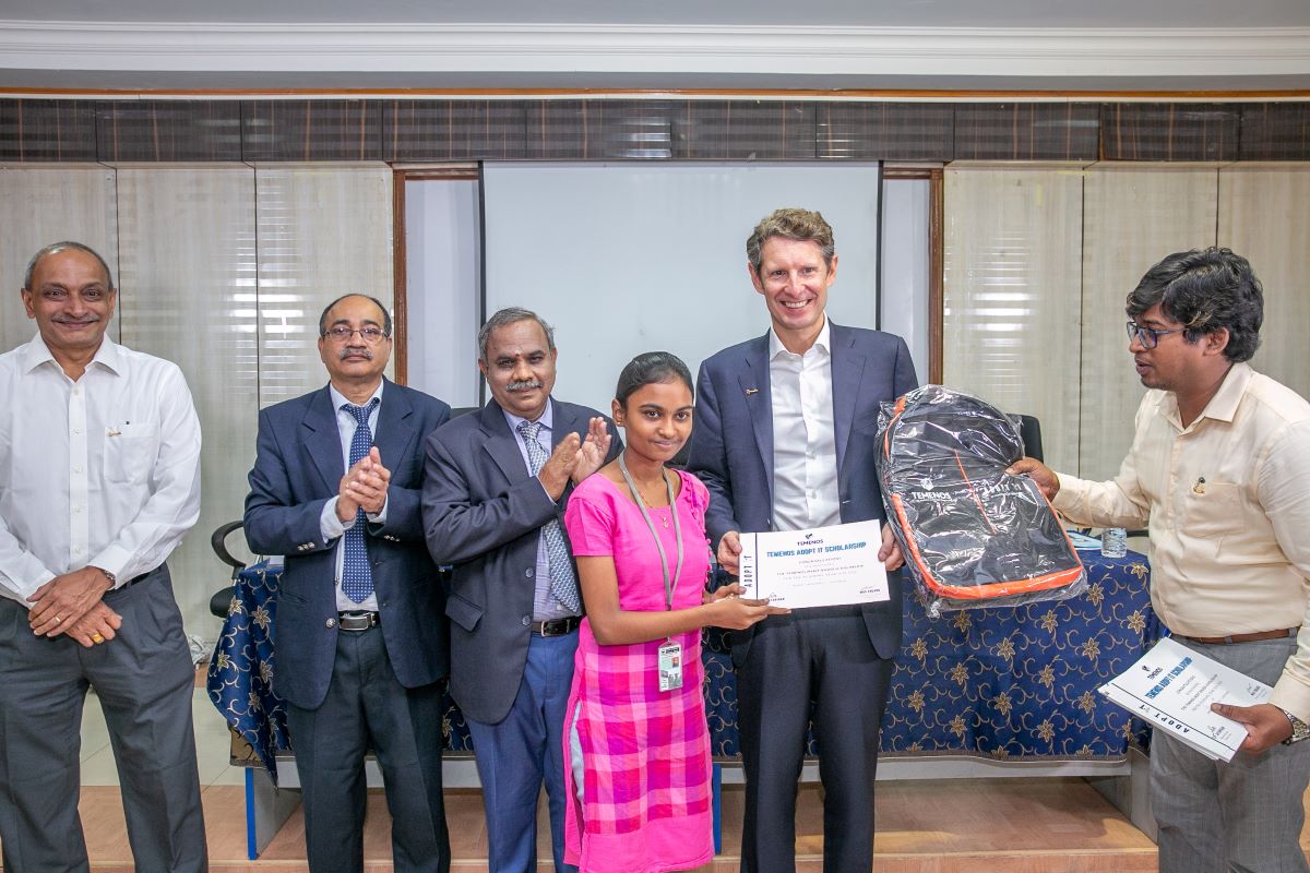 A young girl in a pink dress receives a certificate and backpack, surrounded by smiling, applauding men in suits. The setting is joyful and formal.