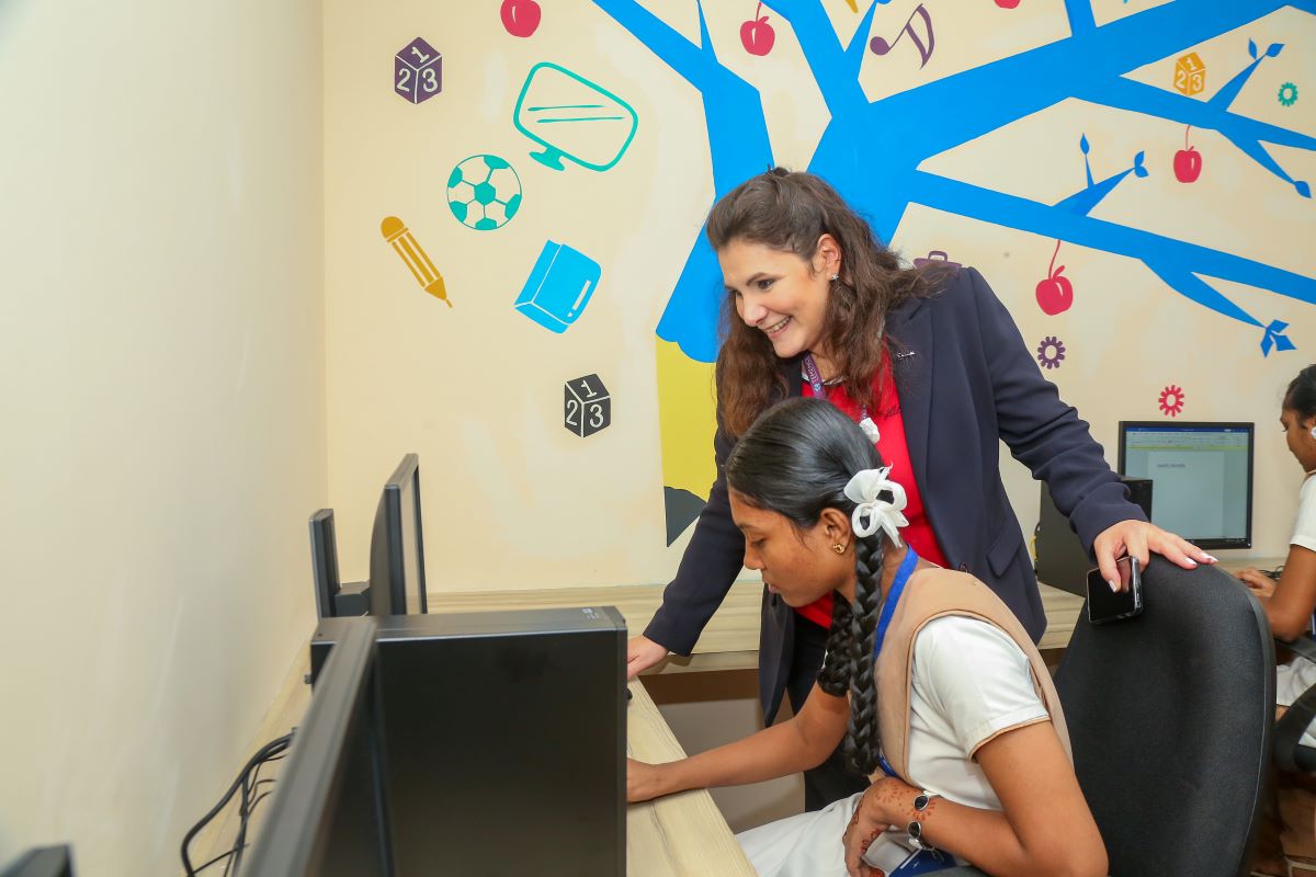 A woman smiles and assists a student working on a computer in a colorful classroom with educational wall art. The mood is supportive and encouraging.