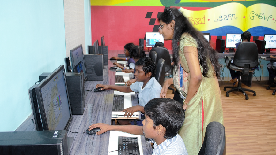 A teacher assists students using computers in a colorful classroom with a wall reading "Read. Learn. Grow." The scene conveys a focused learning atmosphere.