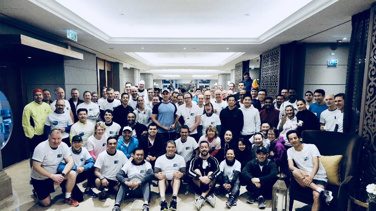 Group photo of diverse people indoors, wearing casual athletic attire and smiling. They're standing in rows in a well-lit hallway, conveying a cheerful and united atmosphere.