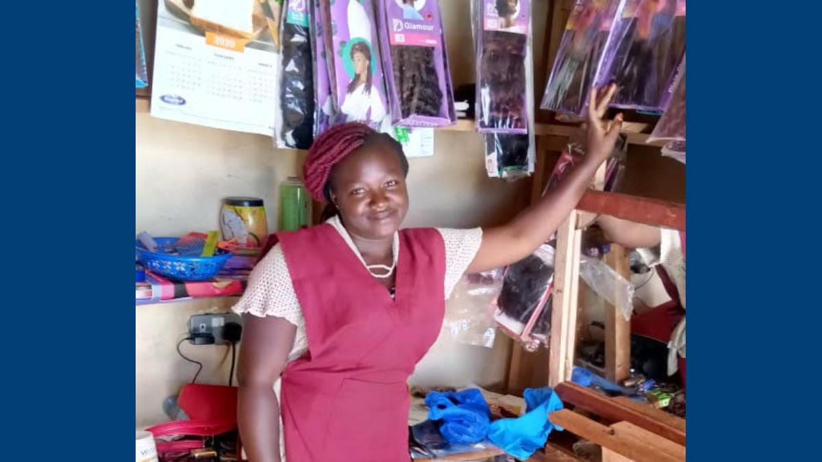 A woman in a maroon dress stands in a store, smiling and pointing at hair extensions. The shop has neatly organized products, conveying a welcoming vibe.