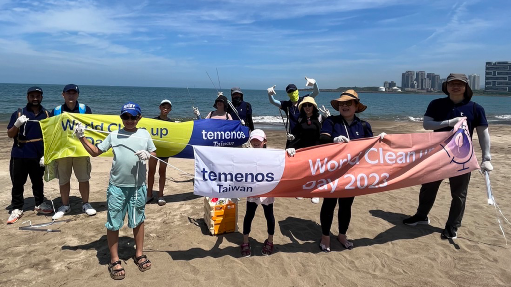 A group on a beach holds "World Cleanup Day 2023" banners, wearing gloves and hats. They're smiling and celebrating under a clear blue sky by the ocean.