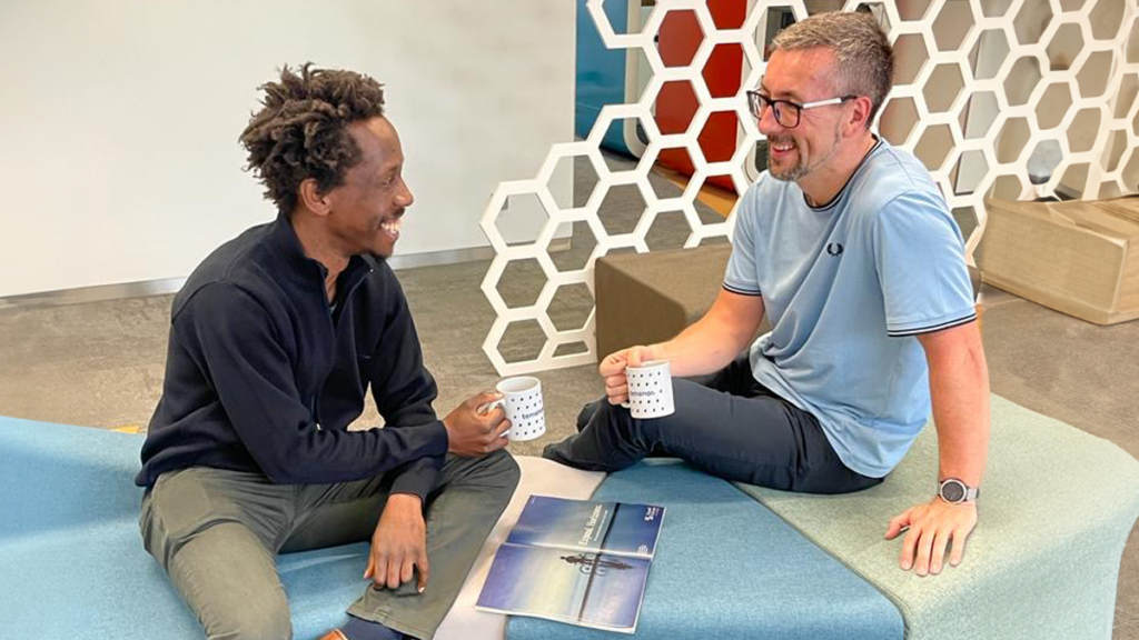 Two men sitting on a blue bench, smiling and holding mugs. In the background is a white hexagonal screen. The atmosphere is friendly and relaxed.