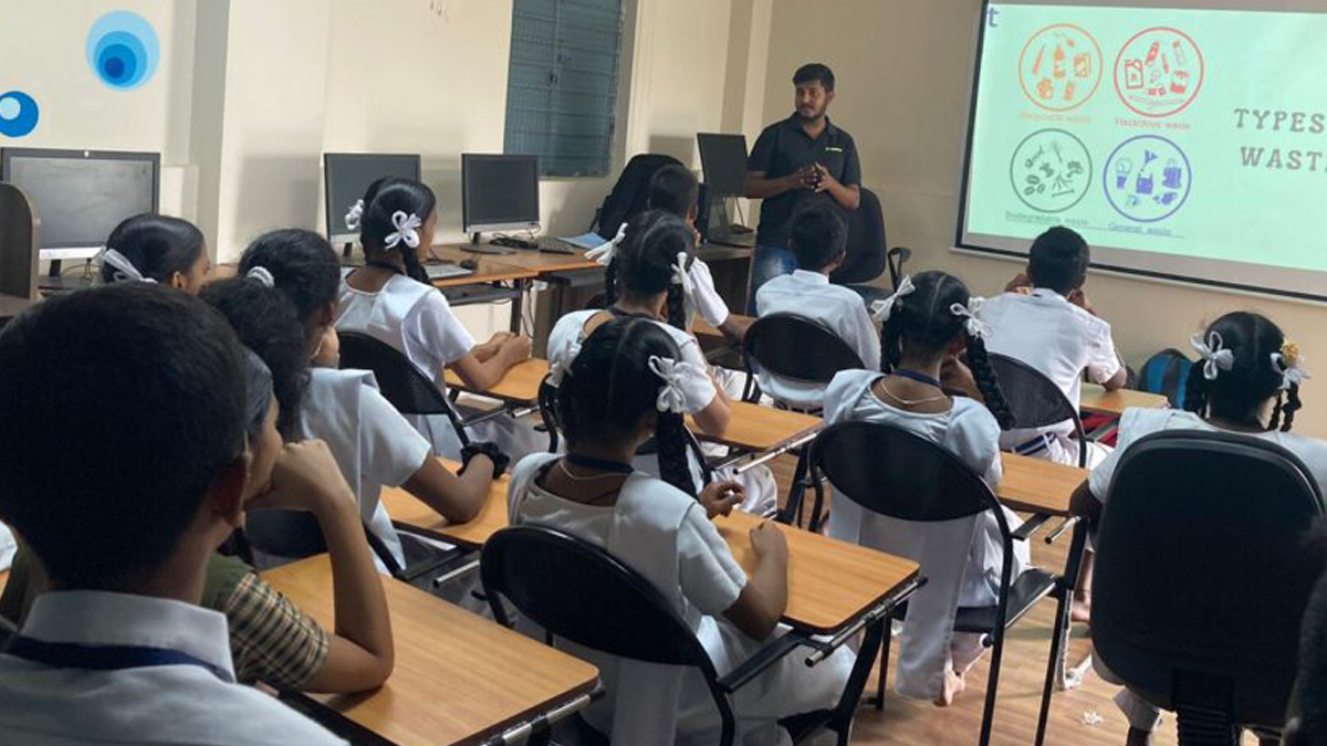 A classroom setting with a male instructor presenting on waste types. Several students in uniforms sit attentively, facing the projector screen.