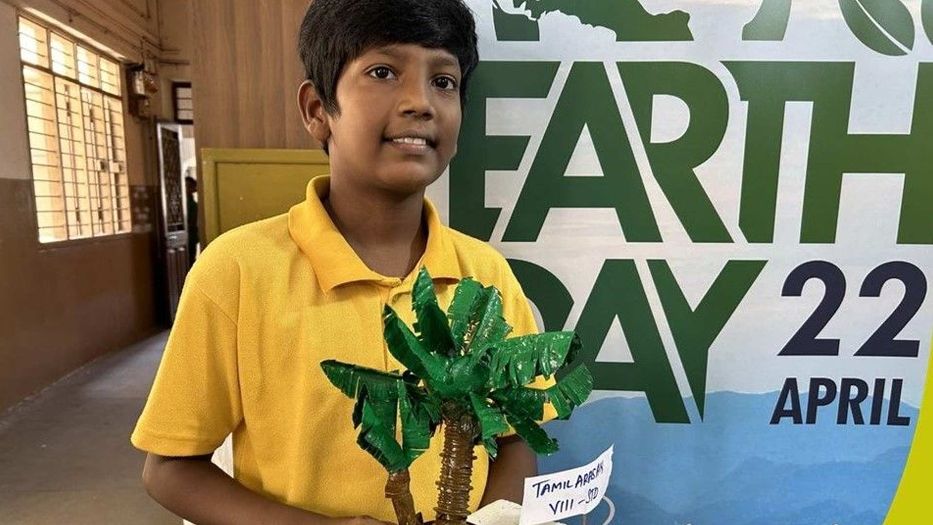 Young boy in a yellow shirt holds a model of a palm tree. He stands in front of an "Earth Day" banner, conveying a sense of environmental awareness.