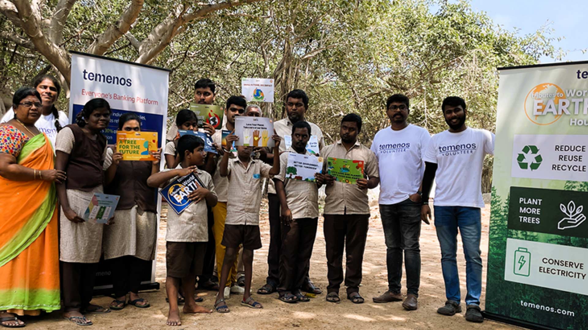 A group of people, including children and adults, stand outdoors holding environmental advocacy signs. Volunteers wear branded shirts. Trees are visible in the background. The mood is positive and educational.