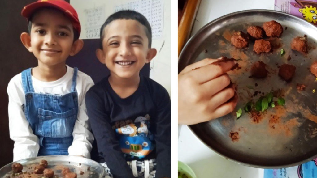 Two smiling children sit together; one wears a red cap and denim overalls, holding a tray with food. Nearby, a hand rolls food on a plate