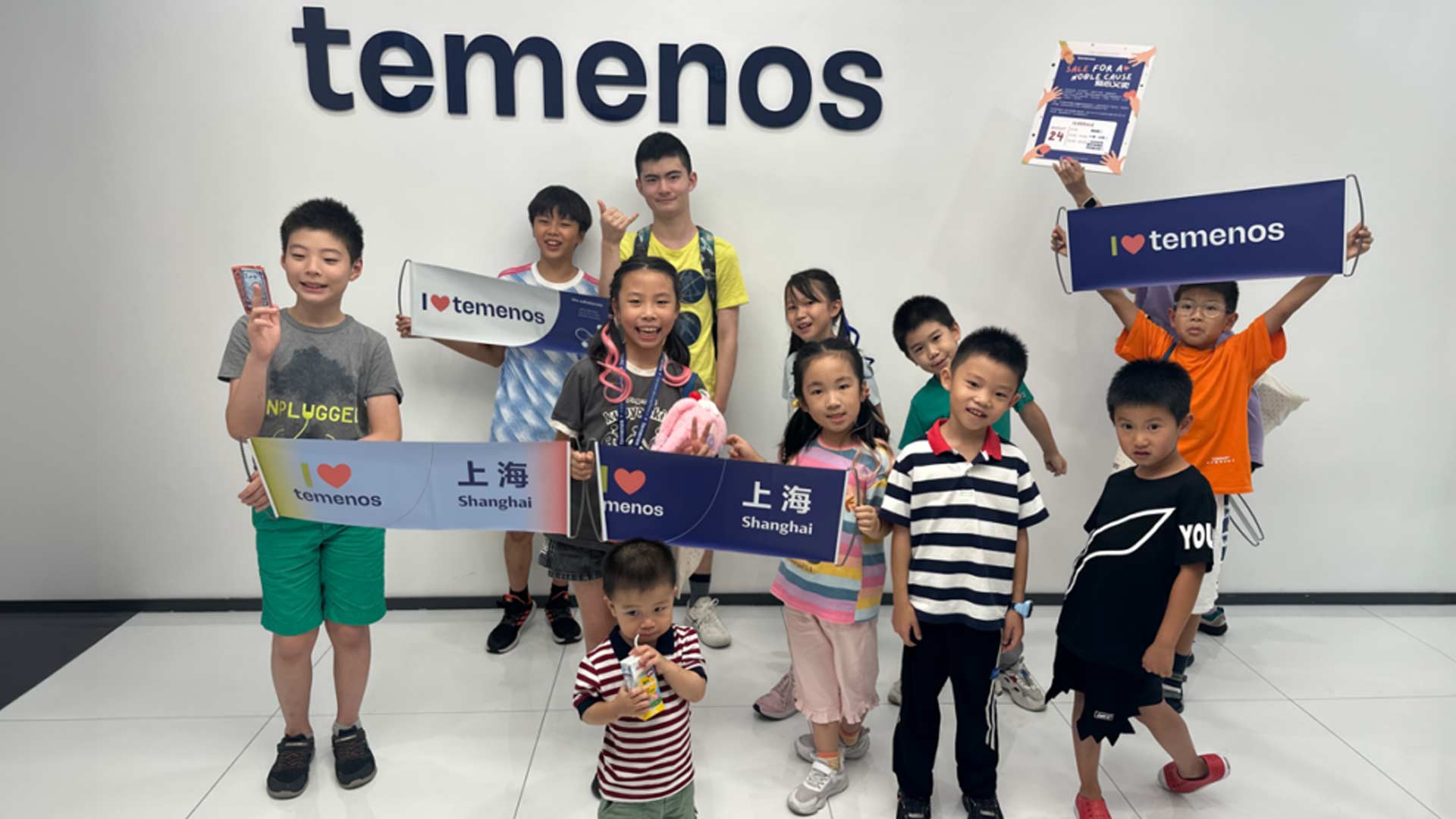 A group of smiling children hold "I love Temenos" and "Shanghai" signs in front of a wall with the Temenos logo. The atmosphere is joyful and lively.