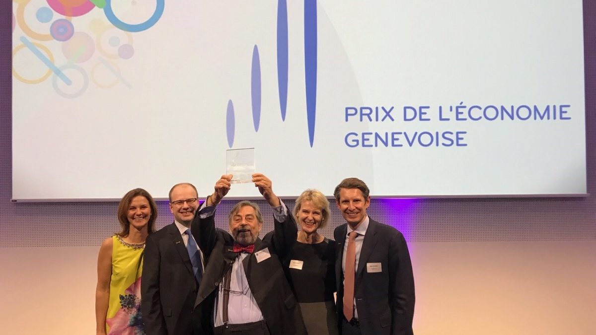 Five people stand smiling in front of a screen displaying "Prix de l'économie genevoise." One person joyfully holds up a trophy. The atmosphere is celebratory.