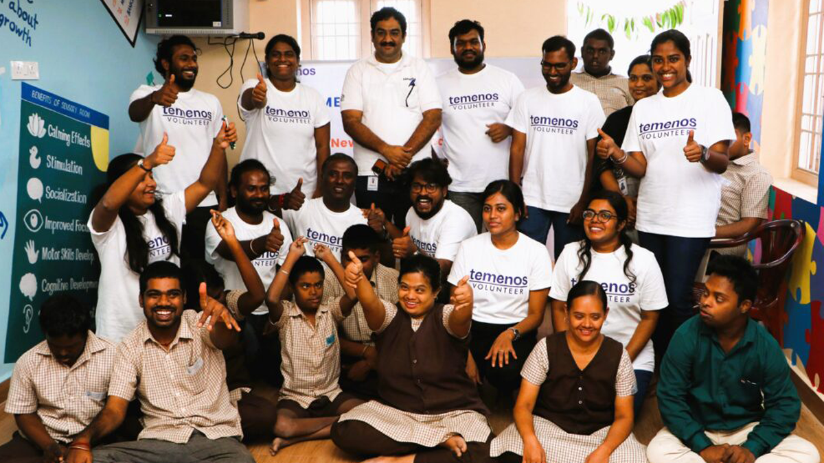 A group of smiling volunteers in white shirts and students in uniforms pose indoors, giving thumbs up. The atmosphere is cheerful and supportive.