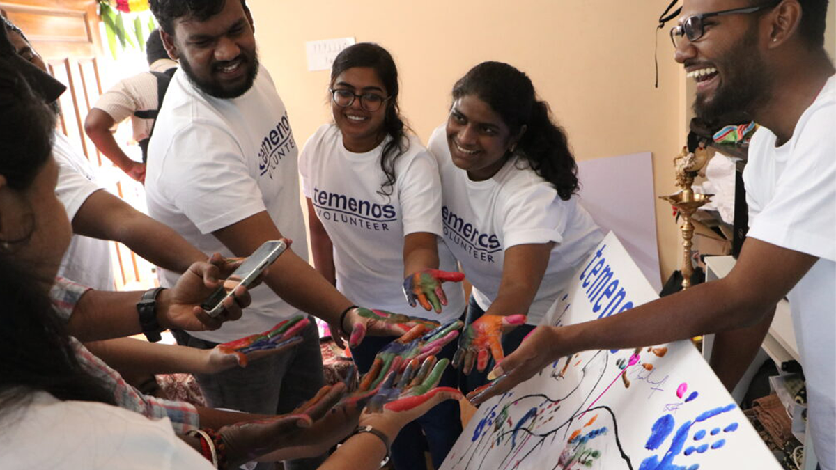 A group of smiling volunteers in white T-shirts with "Temenos Volunteer" printed on them, joyfully display their paint-covered hands around a canvas.