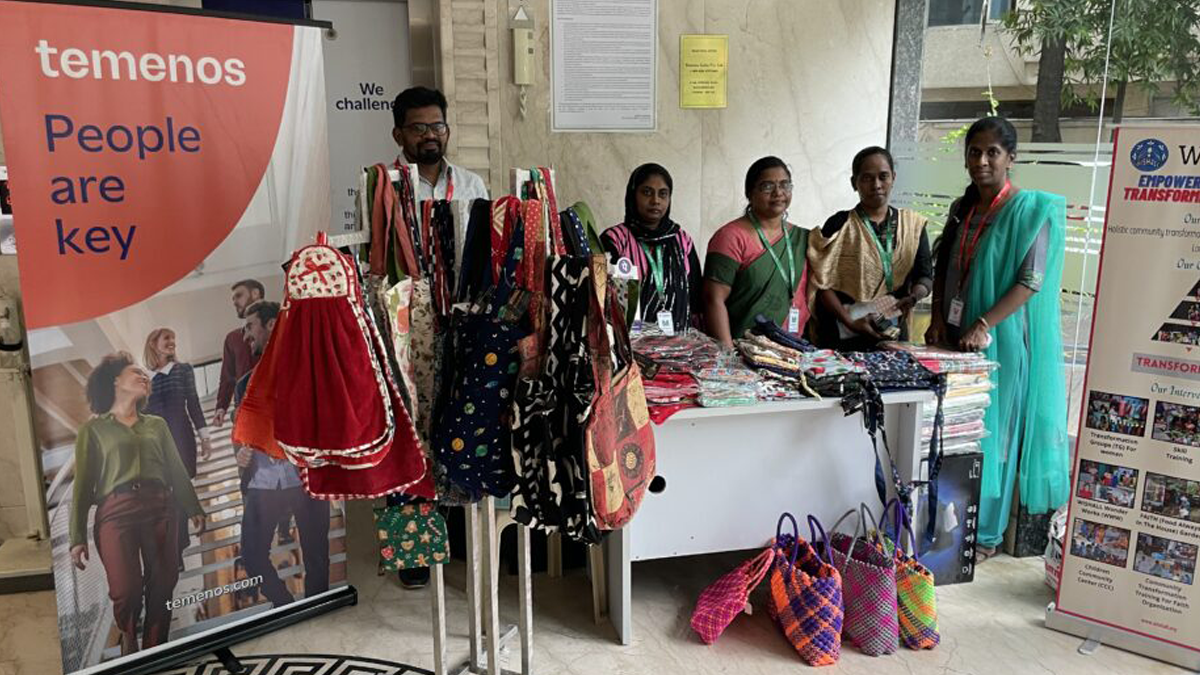 A group of six people stands behind a table displaying colorful fabrics and bags at a market stall. Banners with text "Temenos: People are key" are visible.