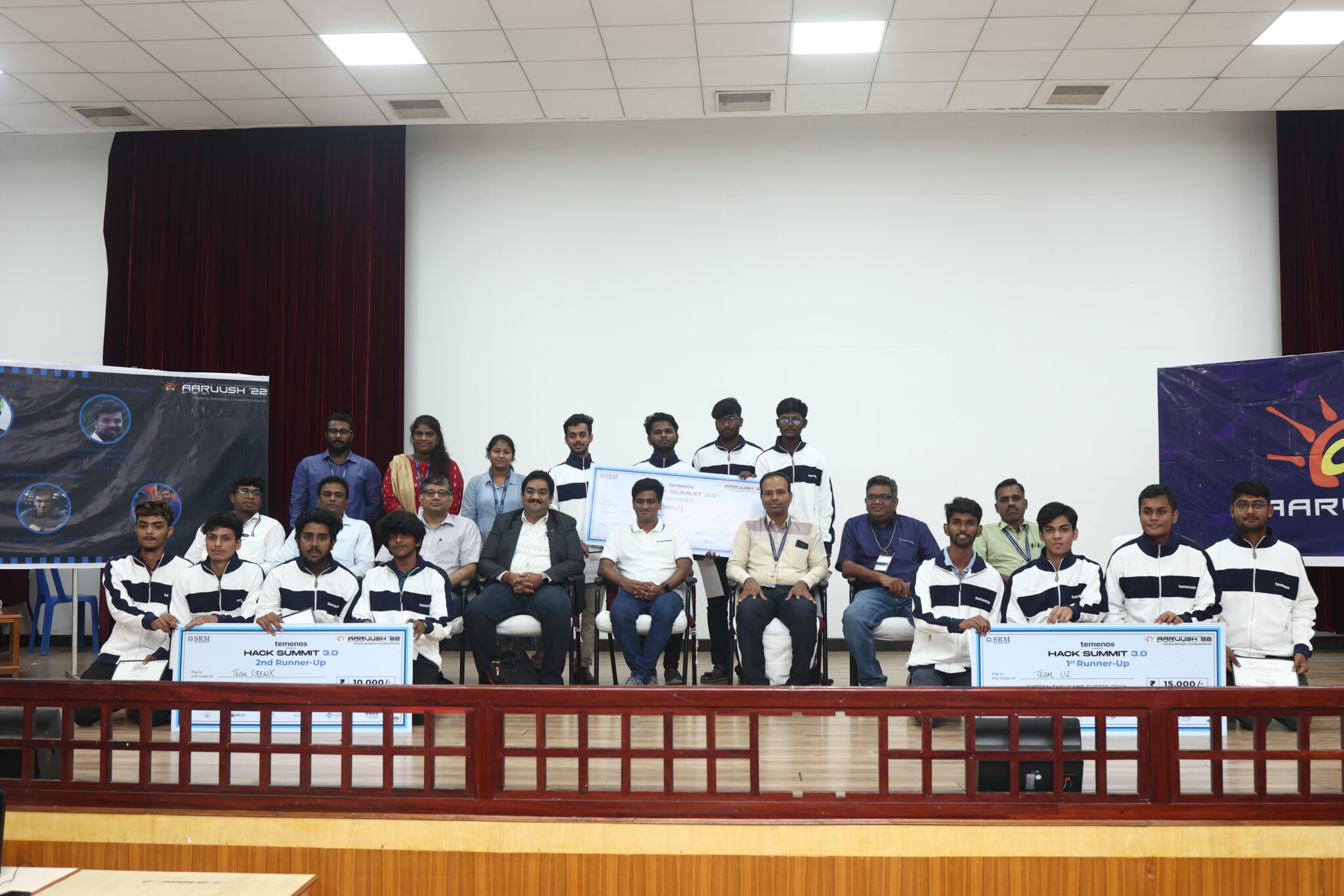 Group photo of a hackathon event, showcasing two teams holding large checks. Participants are smiling and seated in a bright, indoor venue.