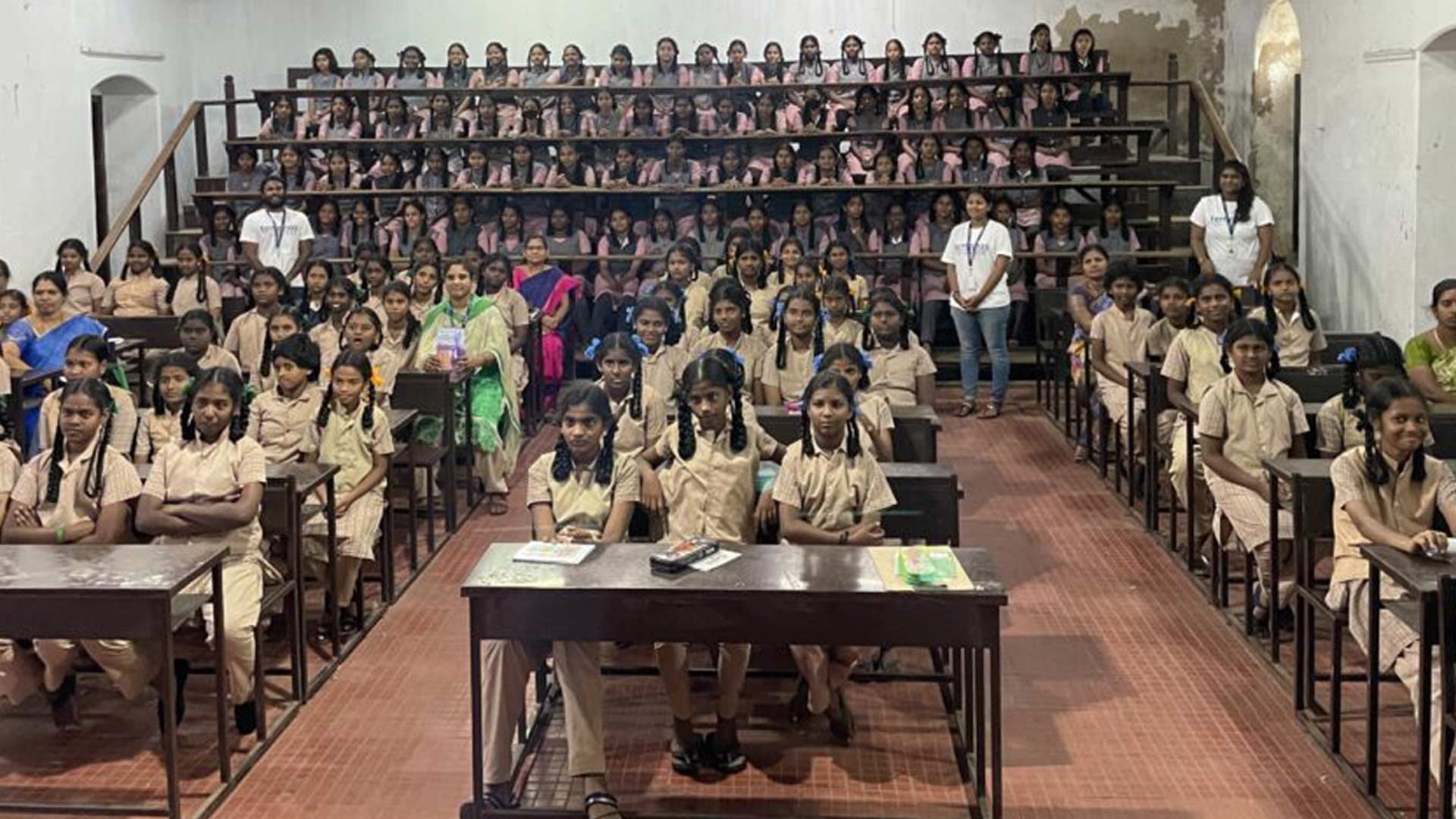 A crowded classroom with rows of young girls in uniforms sitting at desks, and more seated on tiered benches. Two teachers stand on either side, creating a focused, studious atmosphere.