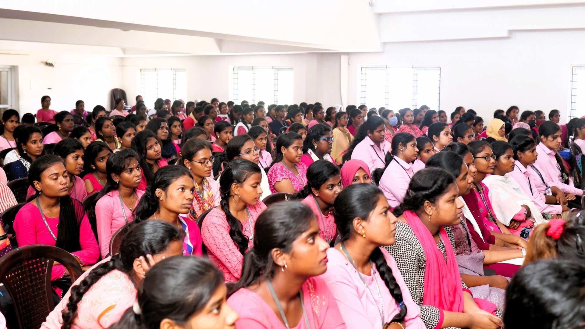 A large group of women in pink attire sit attentively in rows in a bright auditorium. The atmosphere is focused and attentive, with white walls and windows in the background.