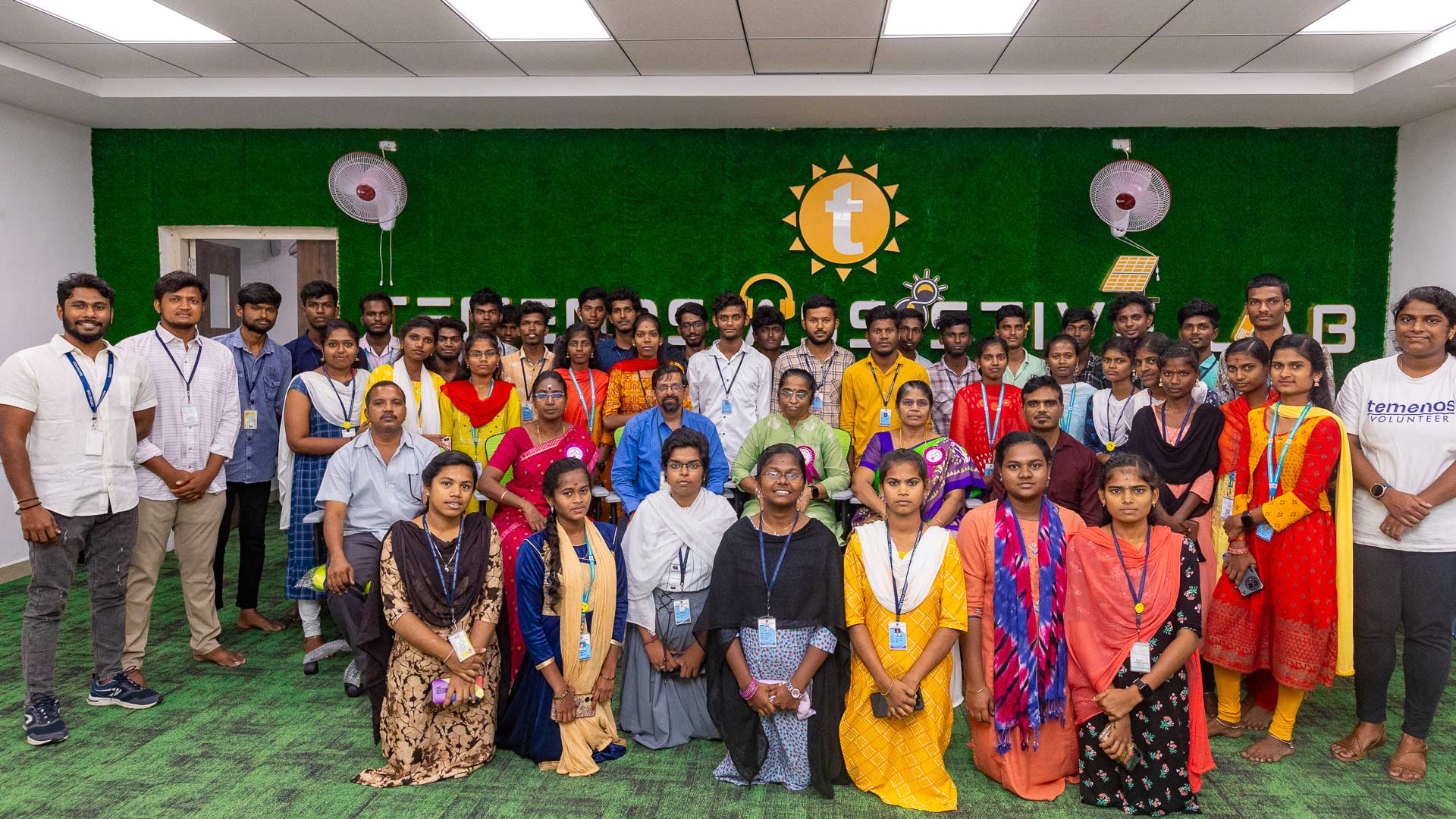 A large group of people, diverse in appearance and attire, pose together indoors against a green wall with a logo. The mood is friendly and inclusive.