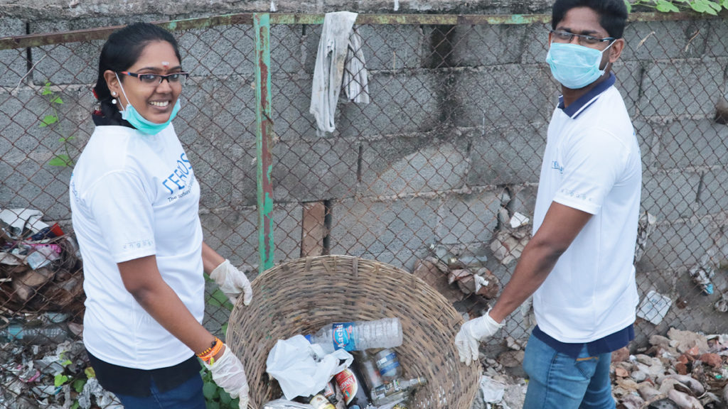 Two people wearing white shirts and masks collect trash into a basket by a chain-link fence. The scene conveys teamwork and environmental care.