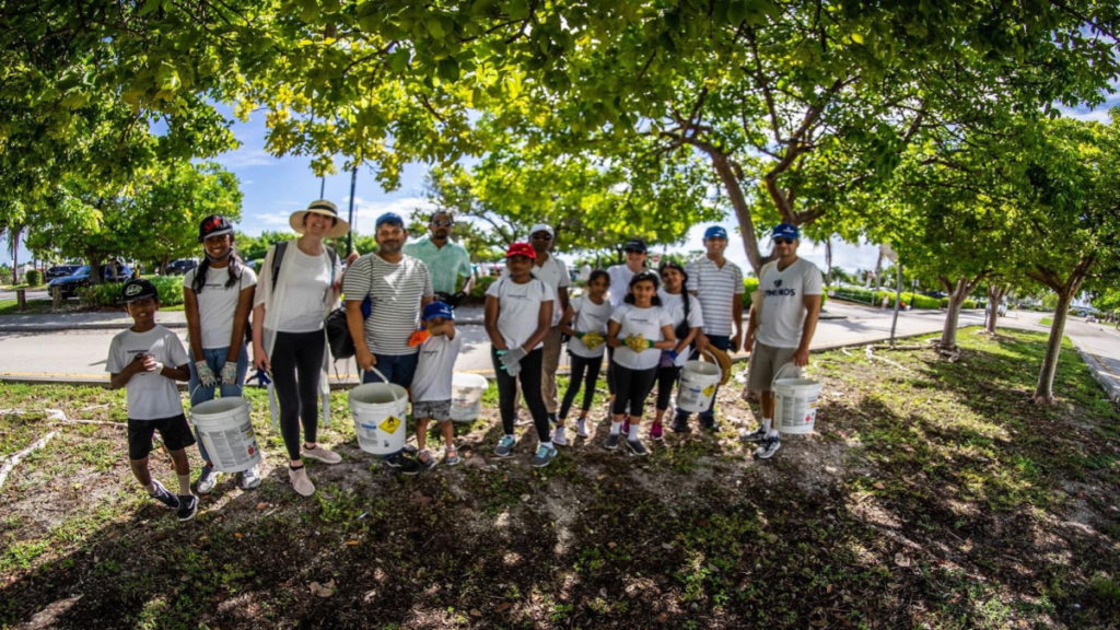 A group of diverse people stands under leafy trees, holding large white buckets. They are smiling, suggesting a community cleanup activity on a sunny day.