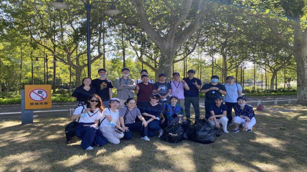 A diverse group of people poses energetically on grass in a sunlit park, surrounded by trees. They smile next to several bags of collected litter.