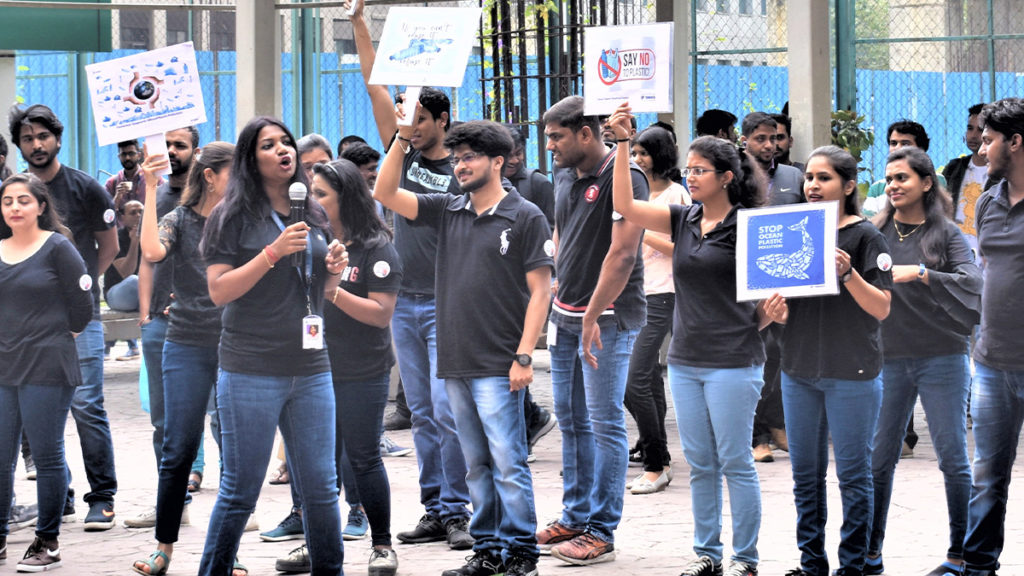 Group of people in black shirts and jeans holding signs during a protest against plastic use. A woman speaks into a microphone, conveying urgency.