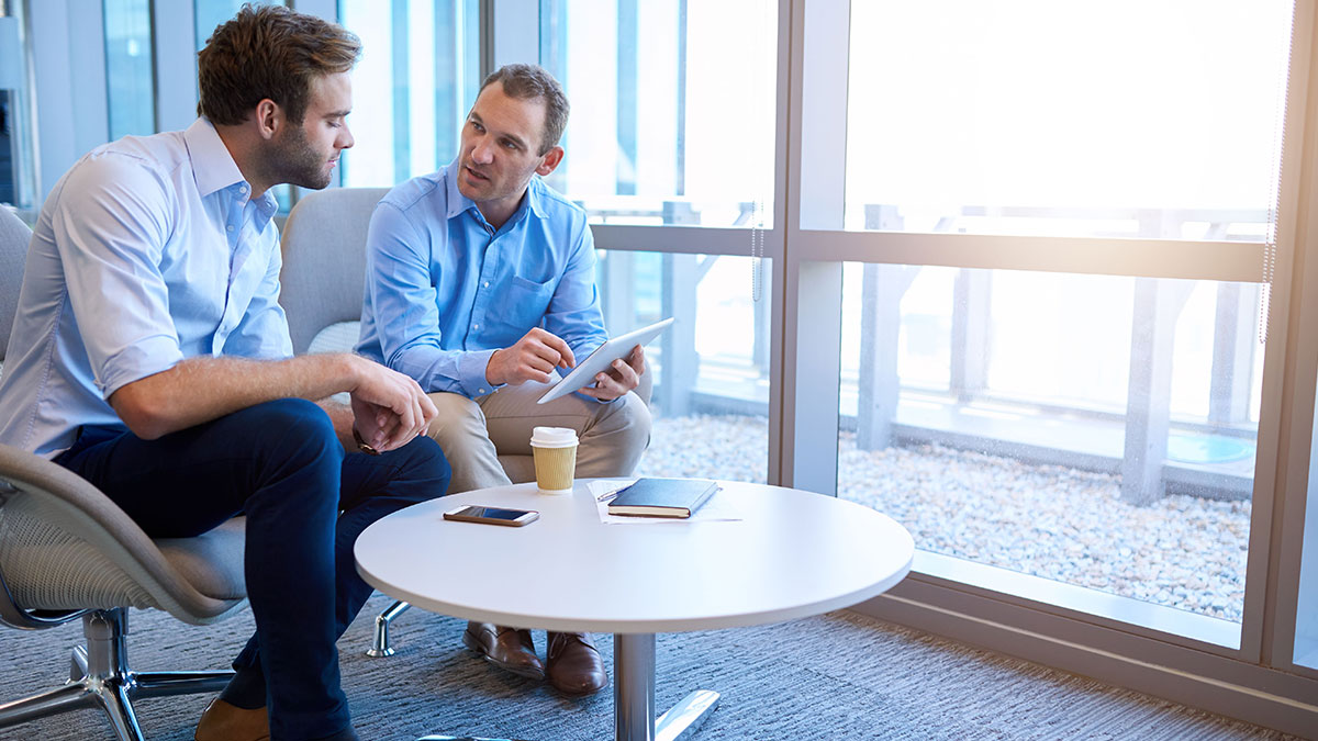 Two men in business attire engage in discussion at a round table. One holds a tablet, and the meeting is held in a bright office with large windows.