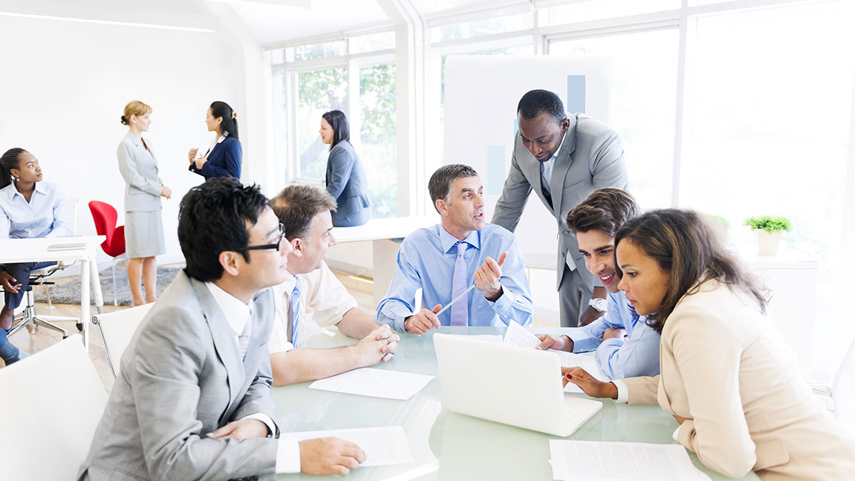 A group of business professionals in formal attire collaborate around a laptop in a bright office. Others converse in the background, creating a focused and engaged atmosphere.