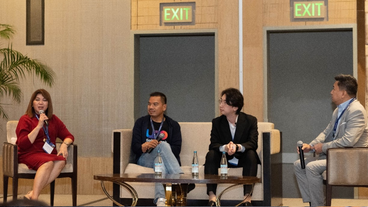 A panel of four people sitting in chairs, one woman speaking into a microphone. They are in a modern conference room with a relaxed atmosphere.