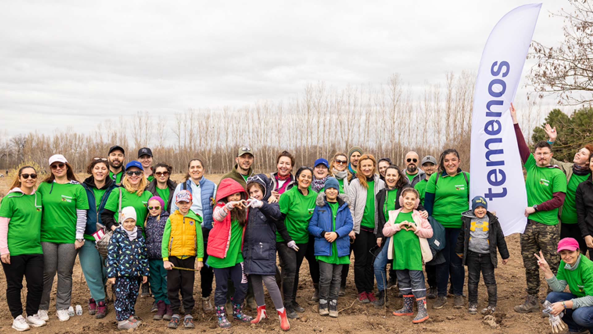 A diverse group of smiling people, including children, in green shirts, stand outdoors on a cloudy day, with a “Temenos” banner signifying teamwork and community.